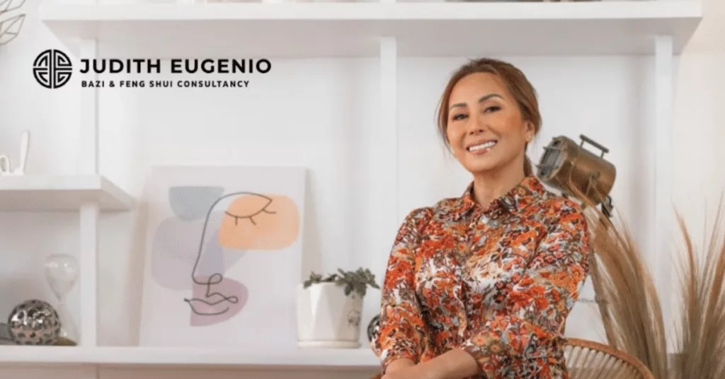 A woman in a floral blouse sits in a room with a minimalist décor that promotes personal growth. Shelves adorned with decorations and a framed abstract face drawing enhance the space's tranquil ambiance, inspired by Feng Shui principles.