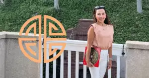 A woman stands on a balcony, smiling. She wears a sleeveless top and holds a red clutch, radiating positive chi energy. A decorative circular symbol adorns the wall on her left. Lush greenery and trees fill the background, enhancing the harmonious atmosphere of her space.