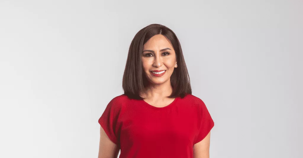 A person with shoulder-length dark hair smiles, wearing a red top against a plain white background.