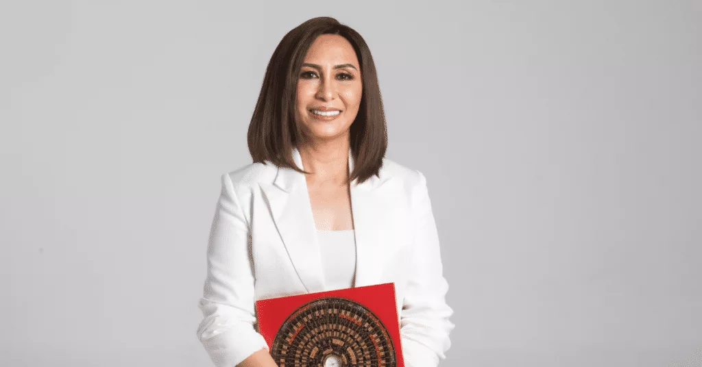 Woman in a white blazer holding a red folder with a decorative cover, standing against a plain background.